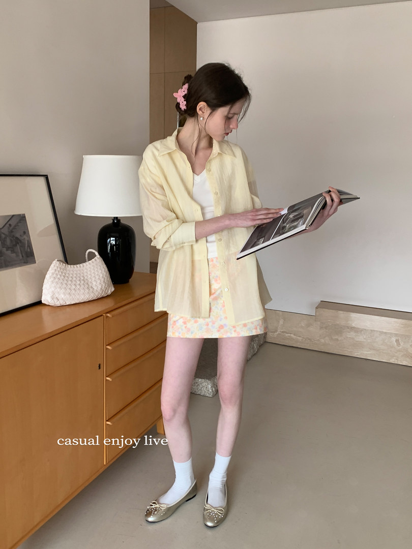 Model wearing a light yellow button-up shirt with a floral mini skirt and flats indoors