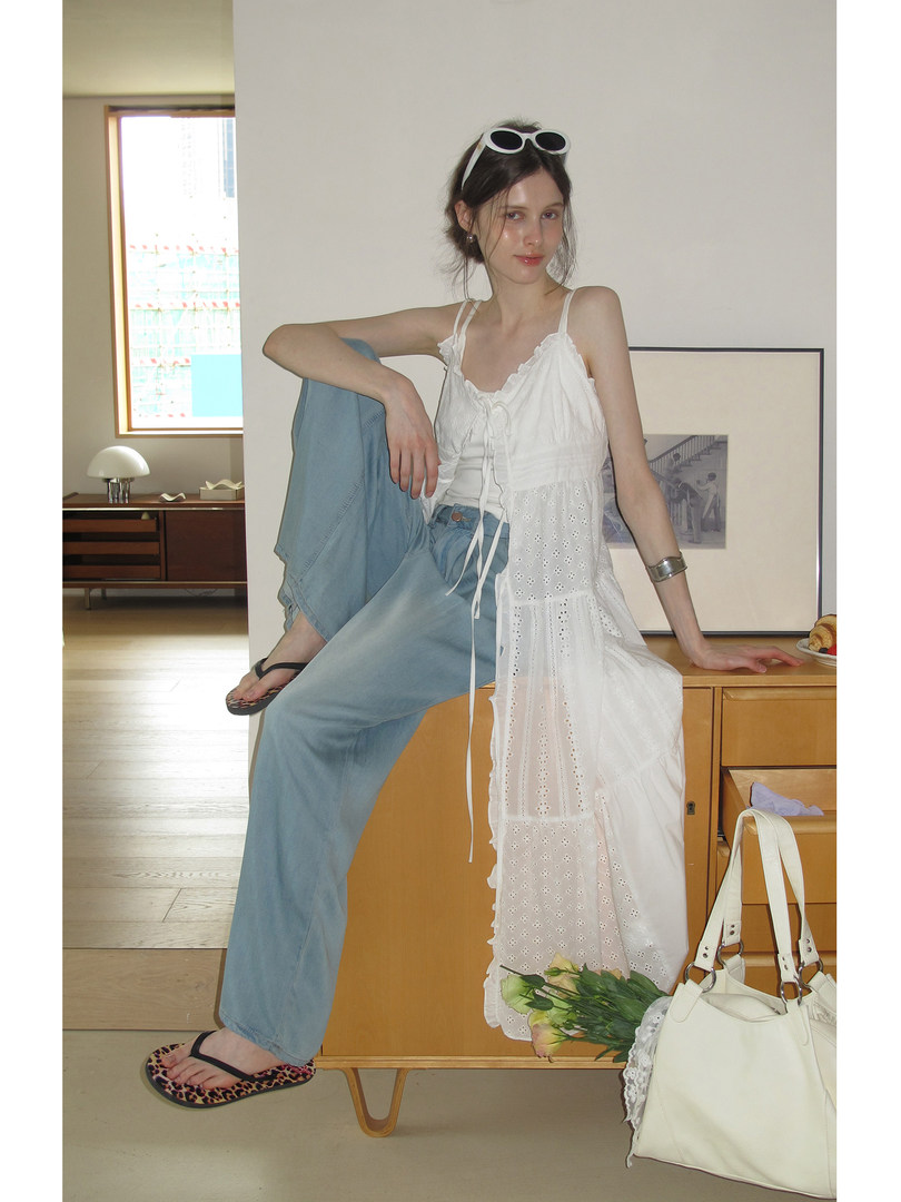 Model wearing a white lace dress layered over blue jeans with a cream handbag and sandals.
