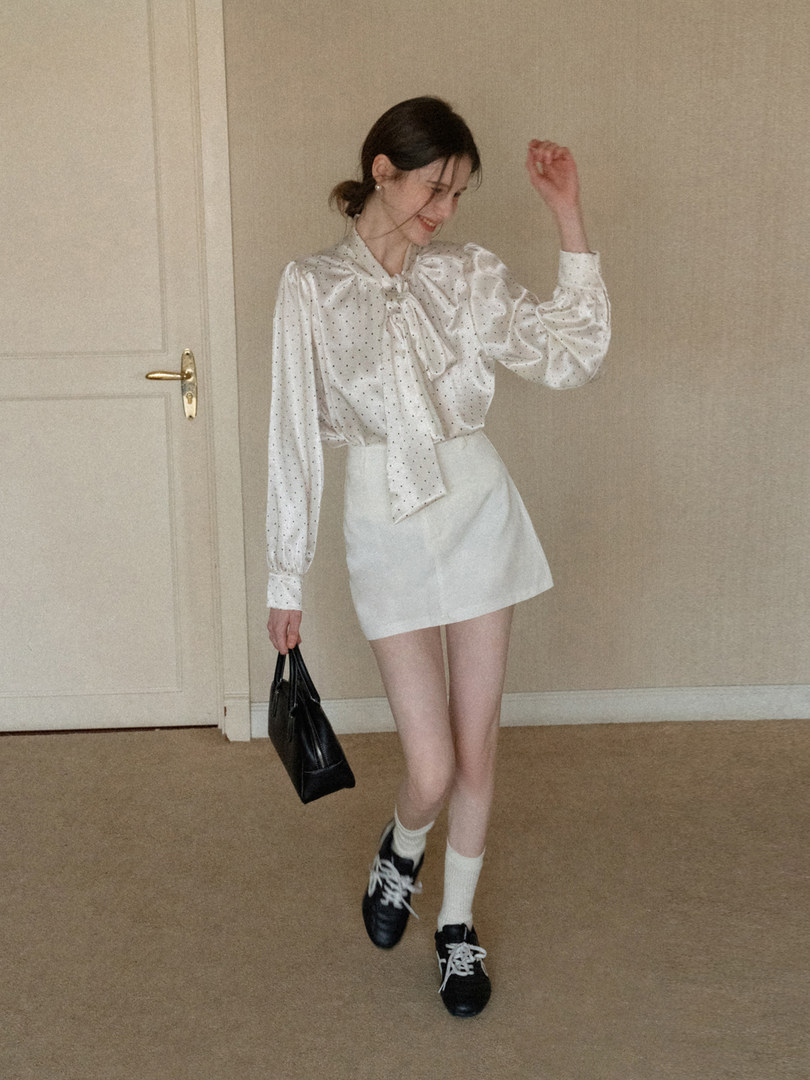 Model wearing a white satin polka-dot blouse with a white mini skirt, black sneakers, and a black handbag