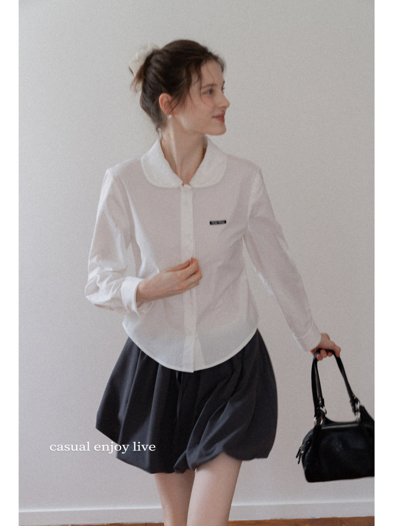 Model wearing a white long-sleeve blouse with a black skirt or shorts and carrying a black handbag