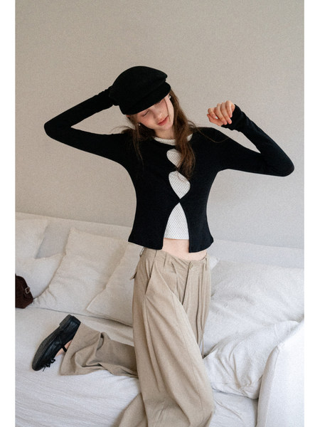 Model wearing a black long-sleeve top with a white patterned cutout layer, beige wide-leg pants, and a black hat