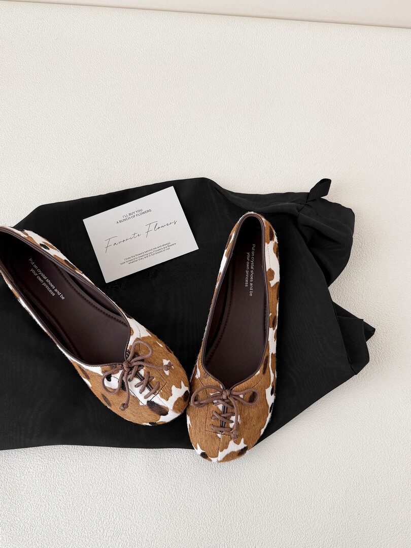 Cow-print lace-up flats displayed on a black garment with a small quote card