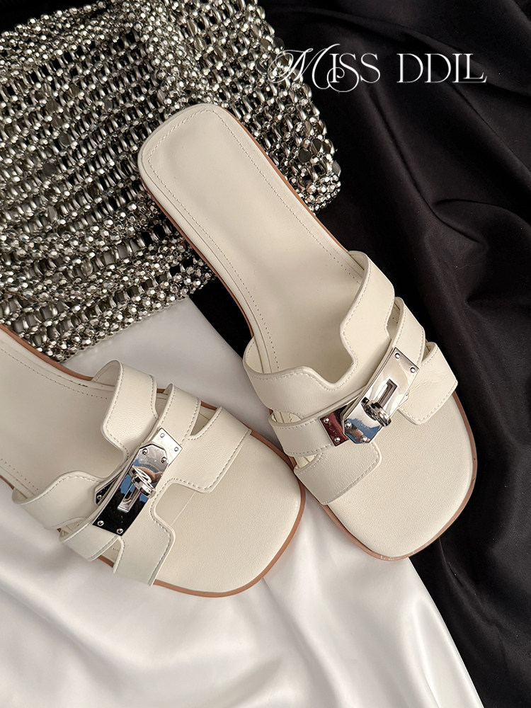 Pair of cream slide sandals with metallic buckle detail on a draped fabric background