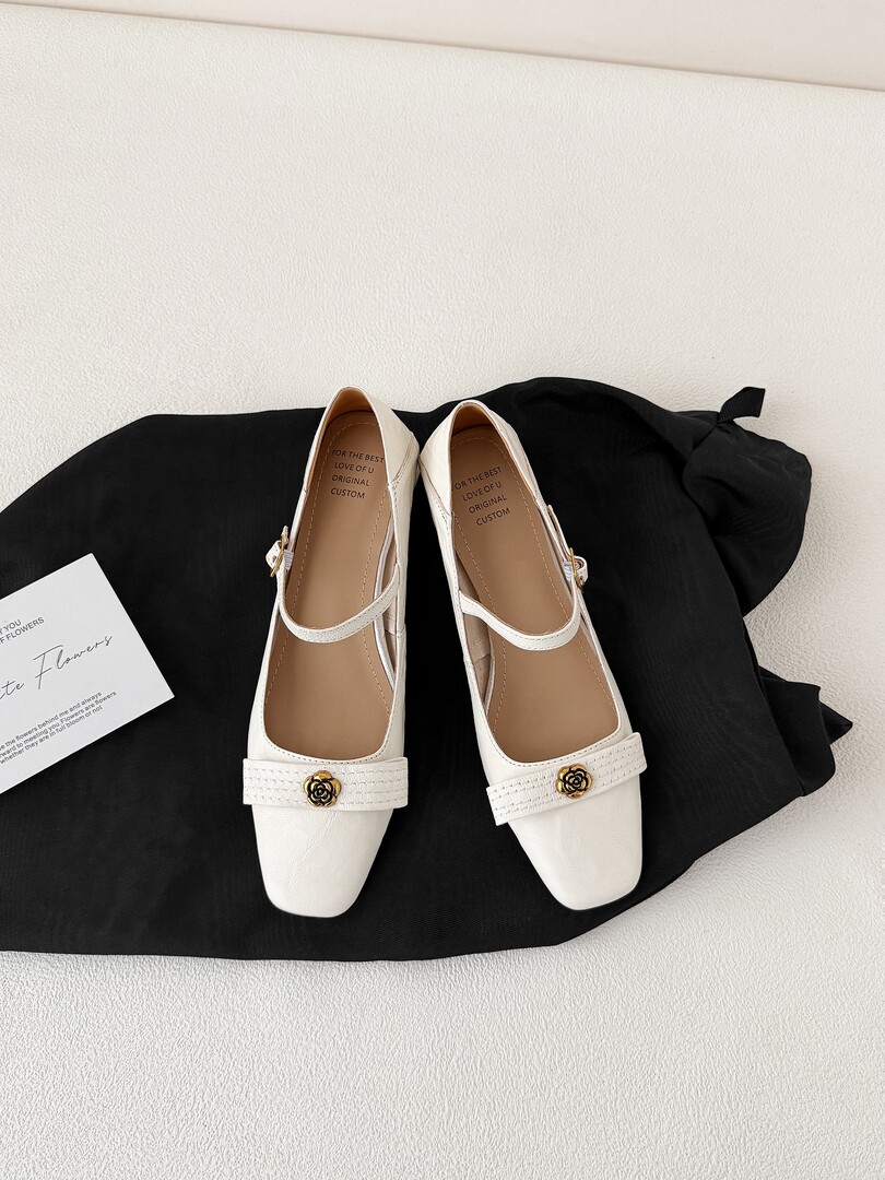 White pointed-toe Mary Jane flats with gold flower detail on a black fabric backdrop