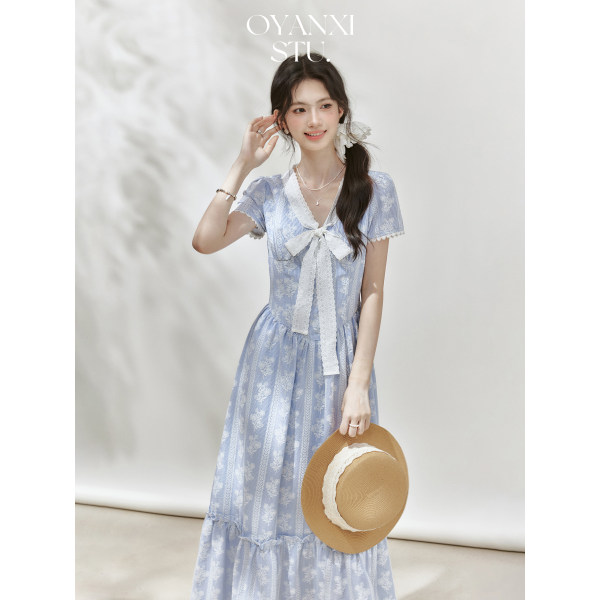 Model wearing a light blue floral lace dress with a bow detail and holding a straw hat