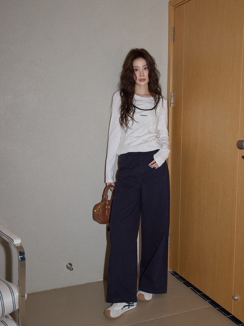 Model wearing a white long-sleeve top with navy wide-leg pants and white sneakers, holding a brown handbag