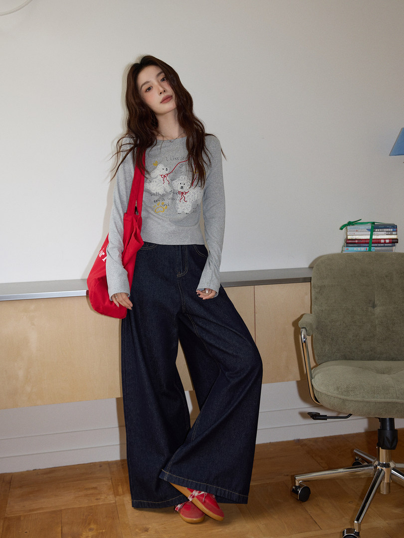 Model wearing a gray graphic long-sleeve top with dark wide-leg jeans and a red tote bag