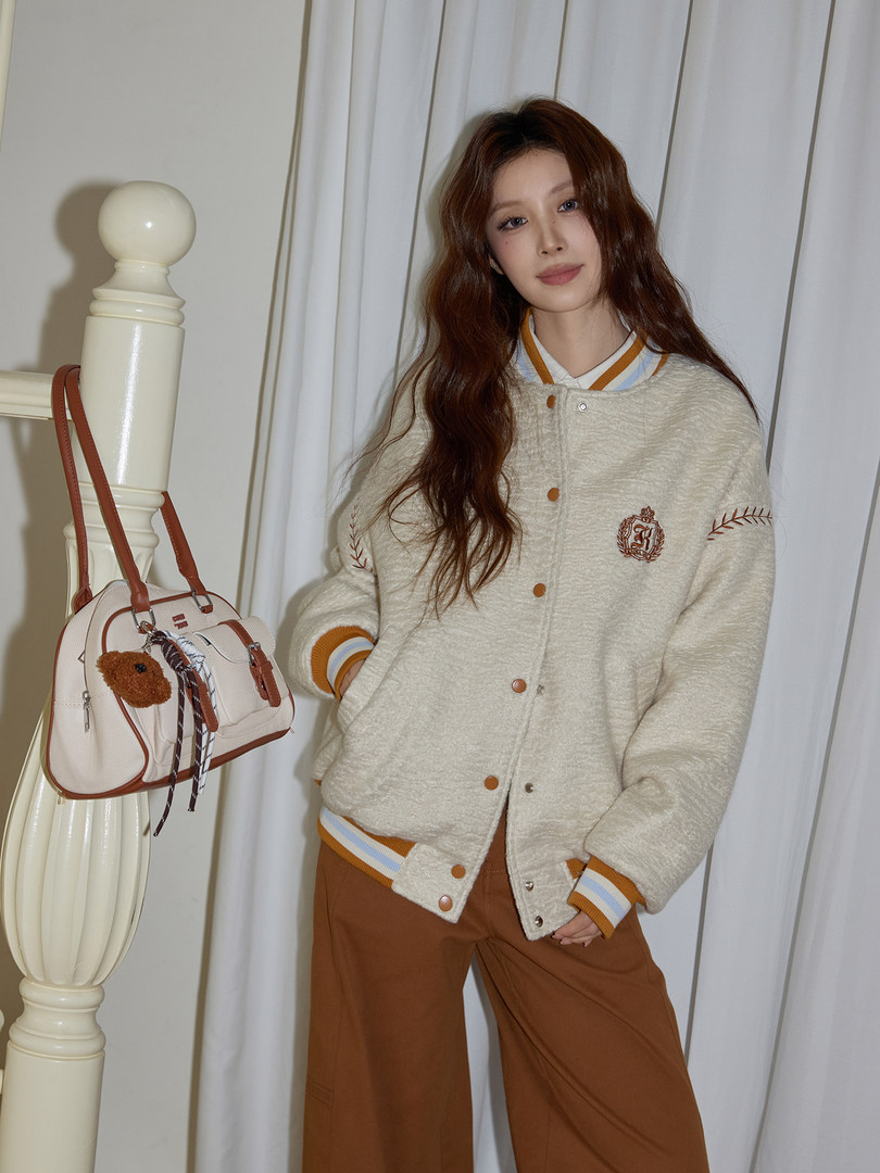 Model wearing a cream varsity-style jacket with brown pants and carrying a beige handbag