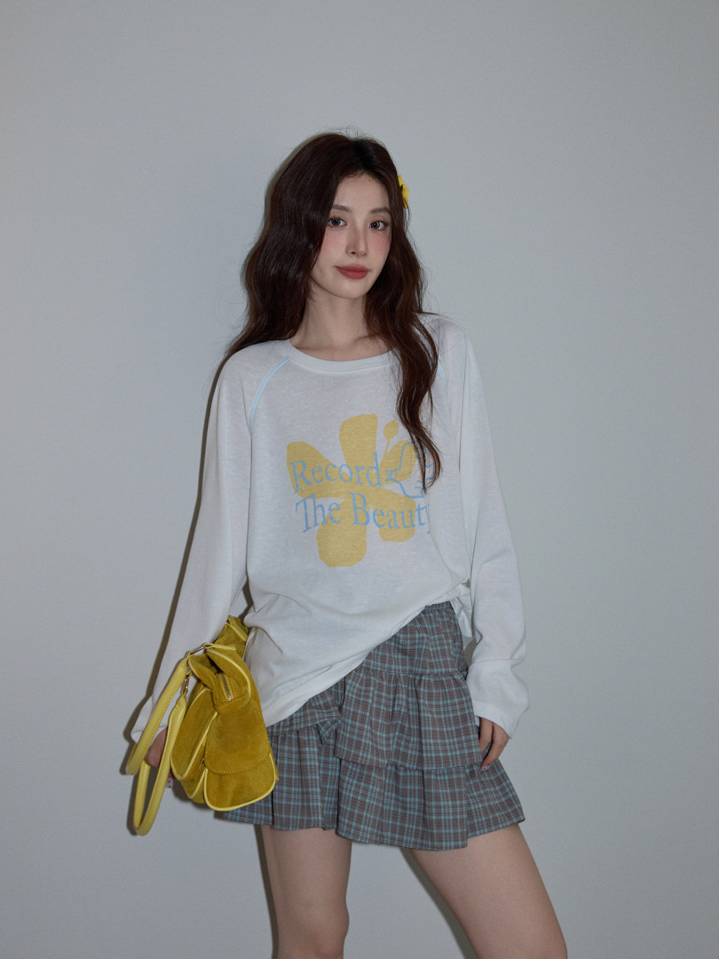 Model wearing a white graphic long-sleeve top with a plaid mini skirt and yellow shoulder bag