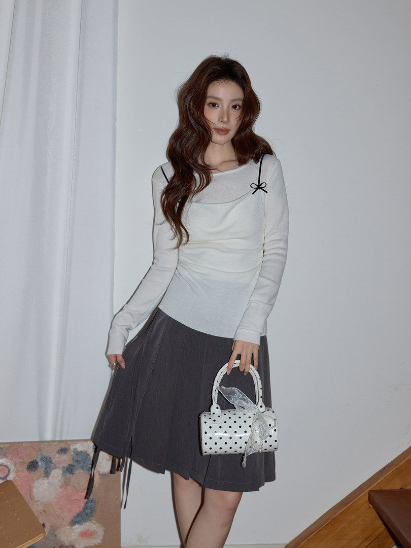 Woman wearing a white long-sleeve top and dark pleated skirt holding a polka dot handbag