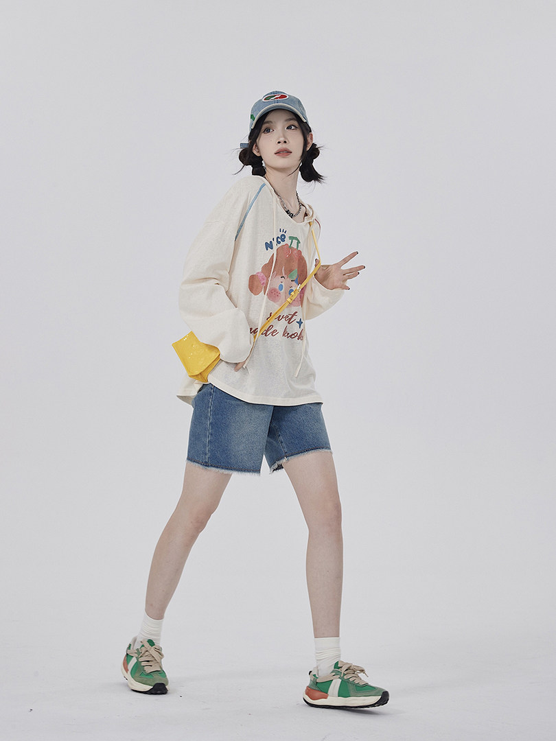 Model wearing an oversized graphic long-sleeve top, denim shorts, and sneakers with a cap and crossbody bag