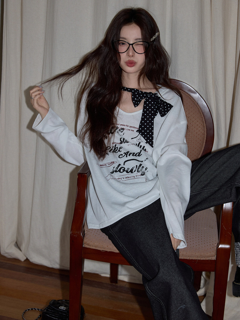 Model wearing a white graphic long-sleeve top with a black polka-dot bow and dark jeans
