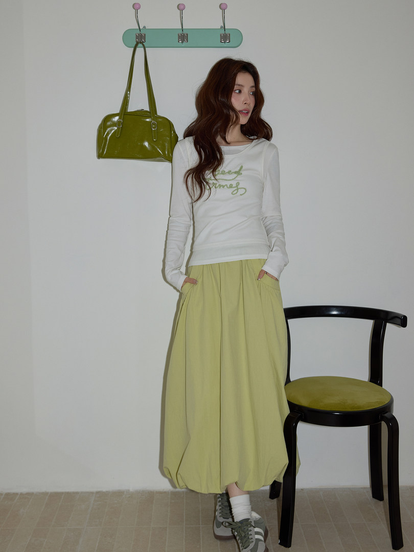 Model wearing a white long-sleeve top with a light yellow skirt and olive green handbag