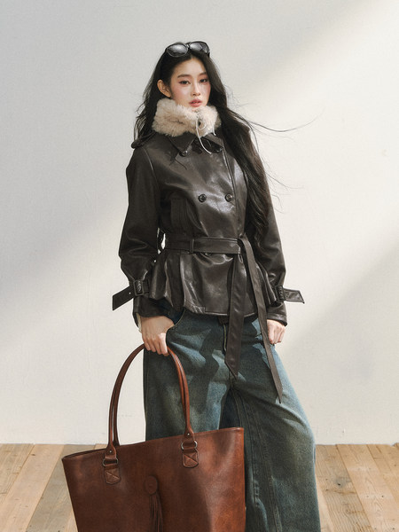 Model wearing a dark leather belted coat with faux fur collar and holding a brown tote bag