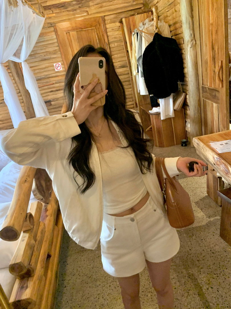 Woman wearing a white casual two-piece outfit with a jacket, tank top, and shorts in a mirror selfie