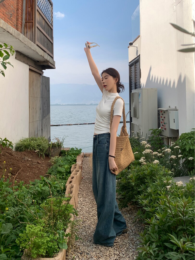 Woman wearing a white fitted top and wide-leg jeans with a woven tote bag outdoors by the water