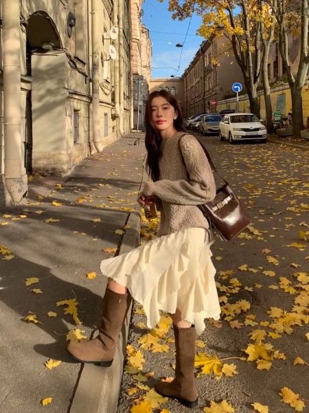 Woman wearing a cozy fall outfit with sweater, skirt, boots, and shoulder bag on a city street