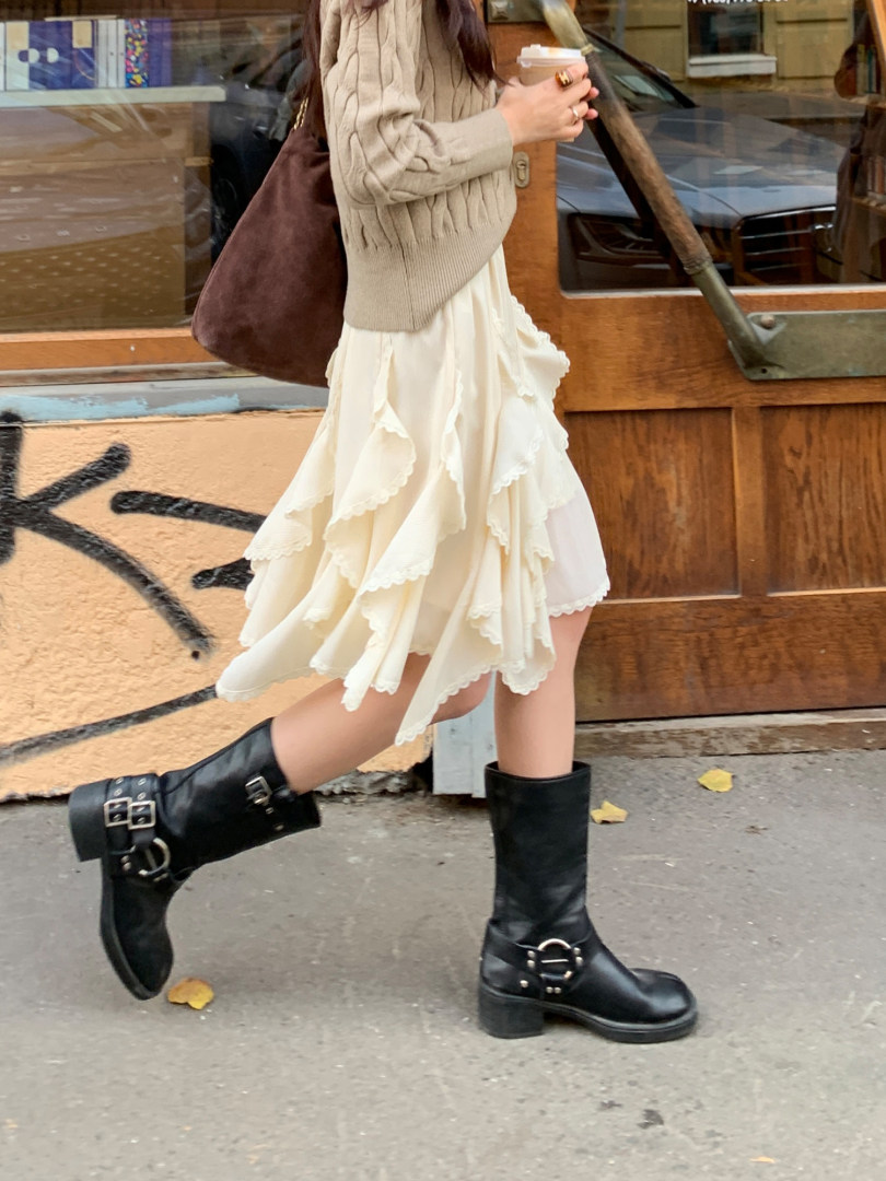 Woman wearing a cream ruffled skirt, beige sweater, and black buckle boots on the street