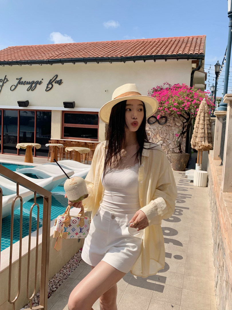 Woman wearing a light yellow sheer shirt, white tank and shorts, straw hat, and designer mini handbag at a poolside resort