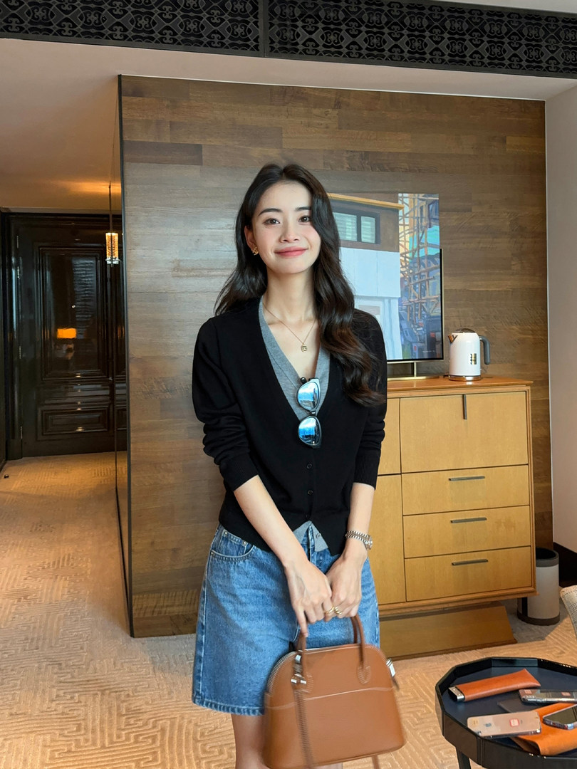 Woman wearing a black cardigan, gray top, denim shorts, and holding a brown handbag indoors