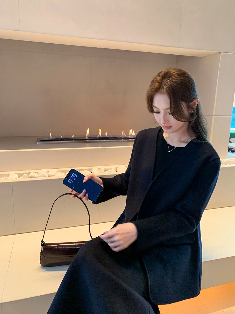 Woman wearing a black long coat and holding a small shoulder bag indoors
