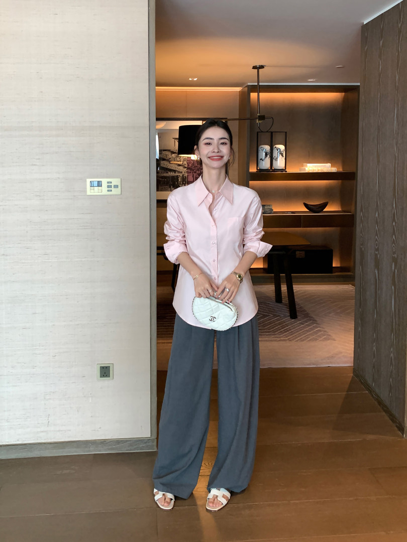 Woman wearing a light pink button-up shirt, wide-leg gray pants, and white sandals while holding a small quilted handbag