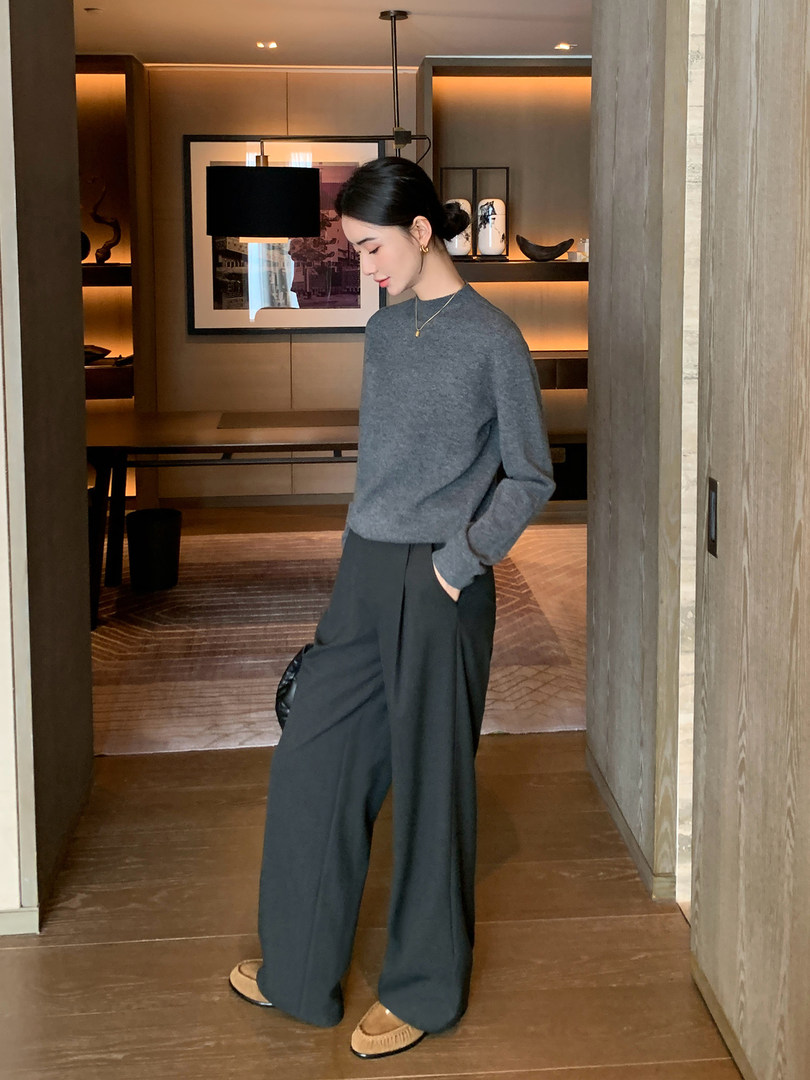 Woman wearing a gray knit sweater with wide-leg dark trousers in a minimalist indoor setting