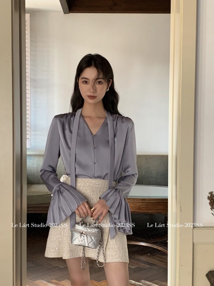 Model wearing a lavender blouse with flared sleeves and a cream textured mini skirt, holding a small silver bag