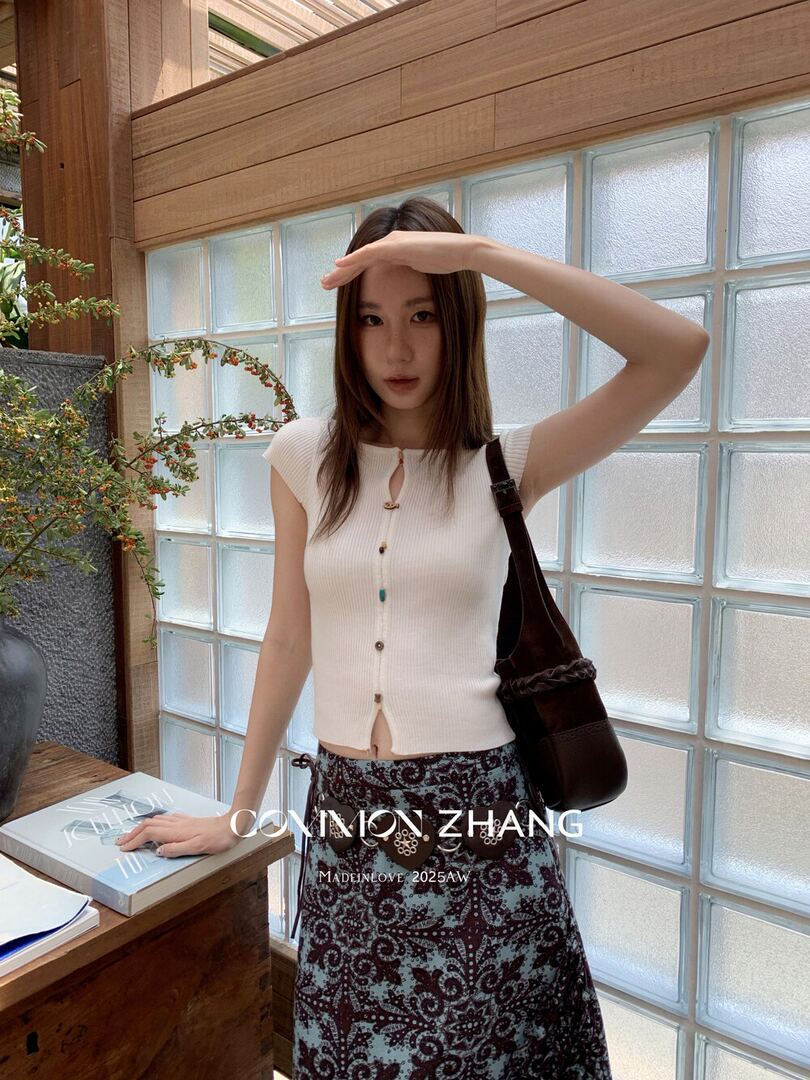 Model wearing a white sleeveless knit top and patterned skirt with a shoulder bag.