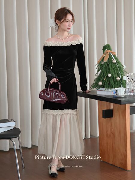 Woman modeling a black off-shoulder dress with sheer hem and ruffle details, holding a burgundy handbag