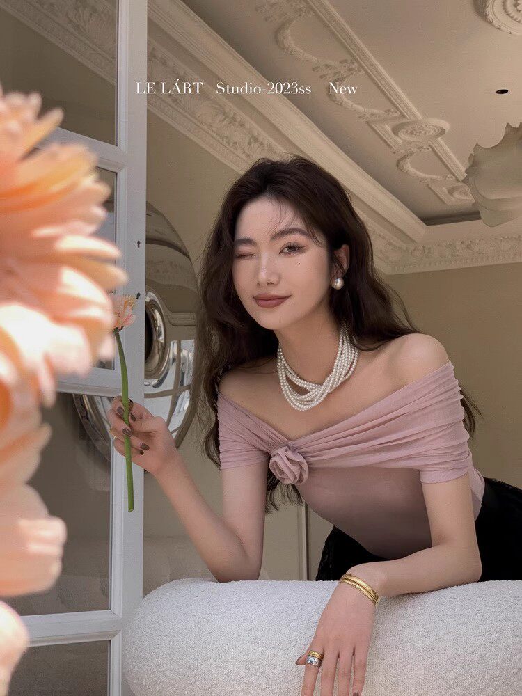 Woman wearing a pink off-shoulder top with pearl necklace in an elegant indoor setting