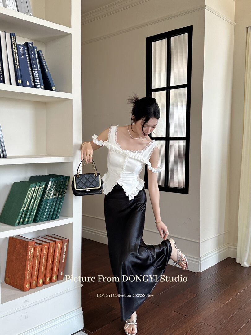 Model wearing a white ruffled off-shoulder top with a black satin skirt and handbag