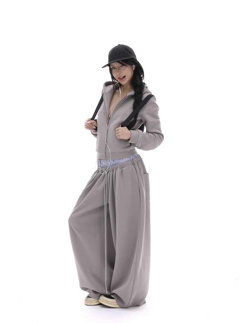 Model wearing a gray zip-up hoodie and wide-leg pants with a black cap on a white background