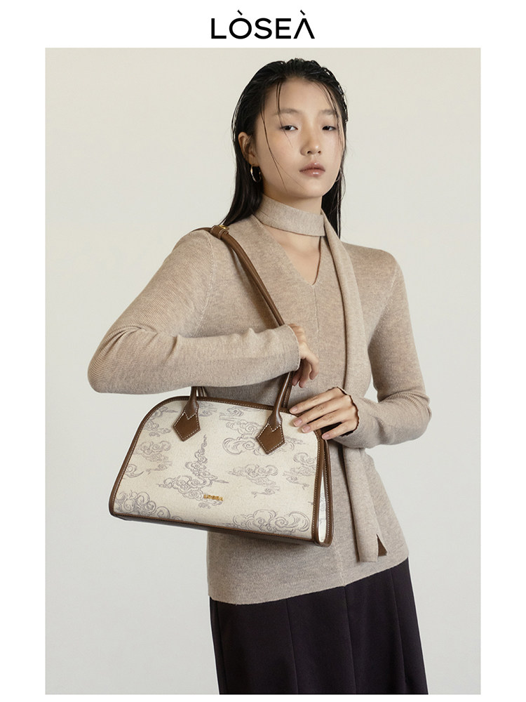 Model holding a beige patterned handbag with brown trim