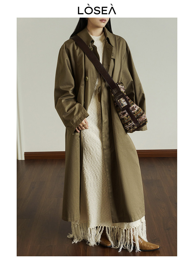 Model wearing an olive trench coat over a cream dress with a crossbody bag