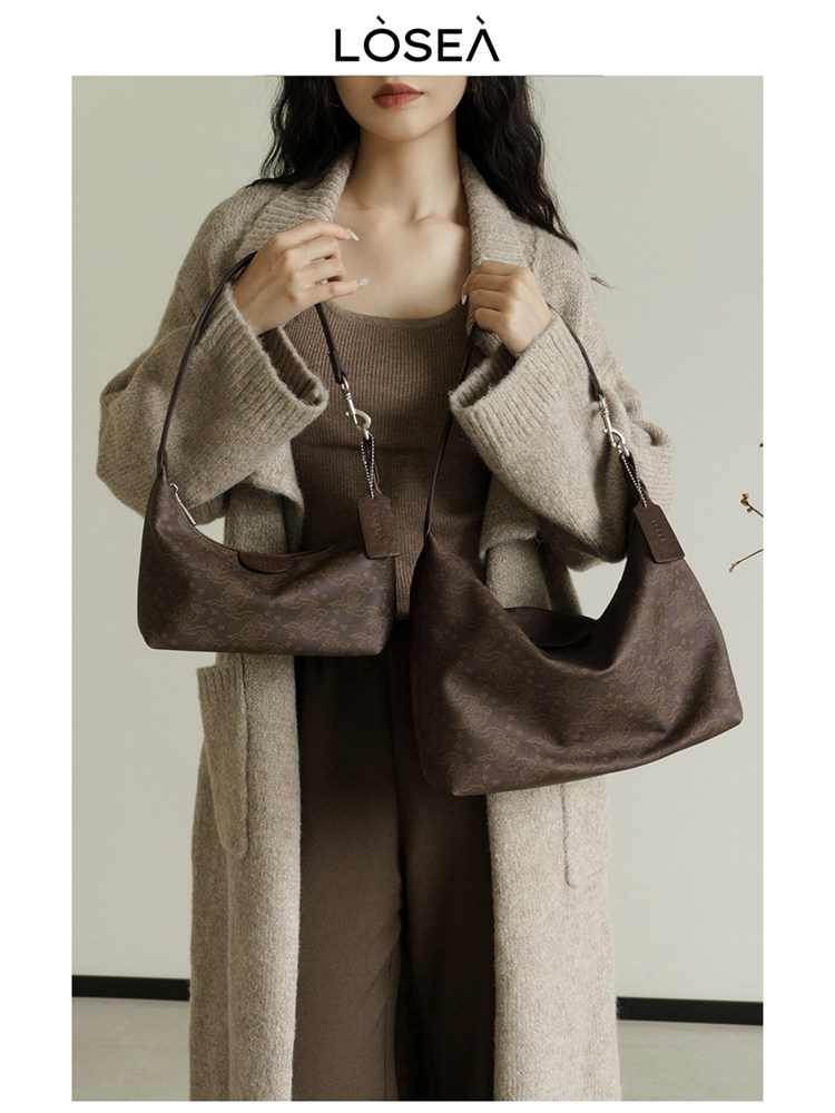 Model holding two brown patterned shoulder bags while wearing a beige knit coat