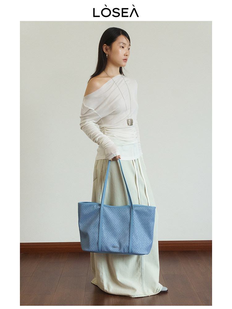 Model holding a light blue tote bag while wearing a white off-shoulder top and long skirt