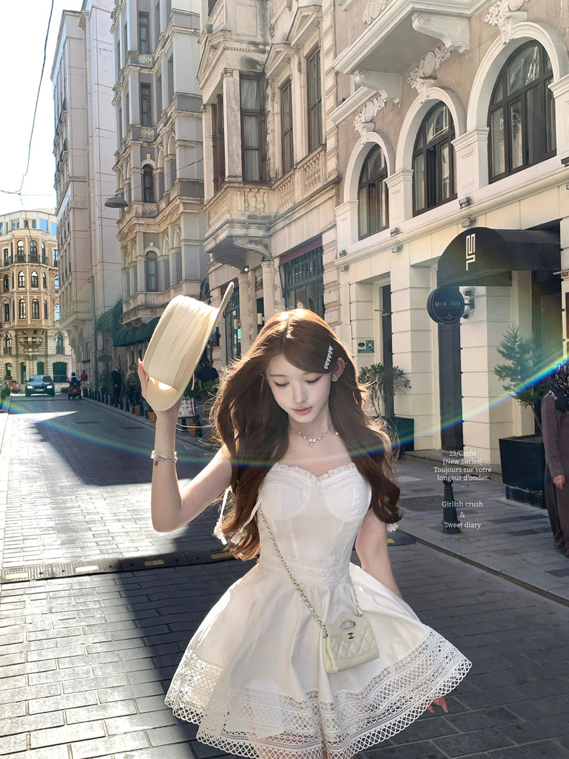 Woman wearing a white lace-trim dress and holding a light-colored hat on a city street
