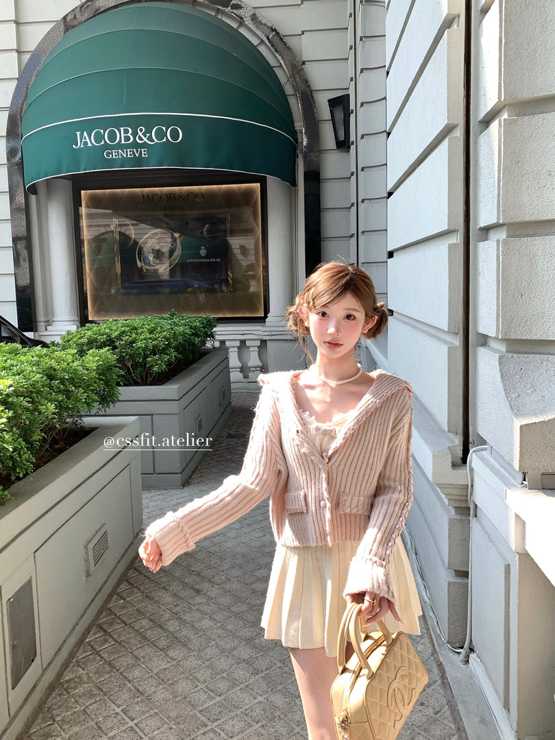 Woman wearing a light pink ribbed cardigan and cream pleated skirt, holding a beige handbag outdoors