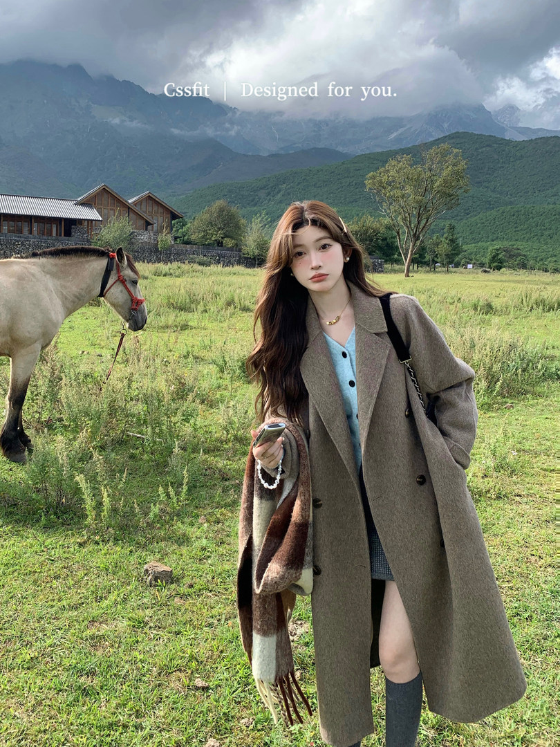 Woman wearing a long brown coat and blue cardigan in a scenic outdoor field