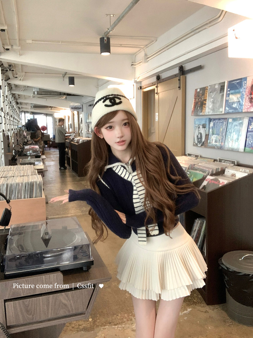 Woman wearing a navy sweater, striped top, white pleated skirt, and cream beanie in a record store