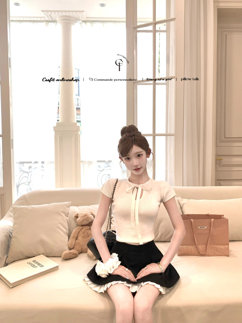 Model wearing a white tied-front top with a black skirt seated on a sofa