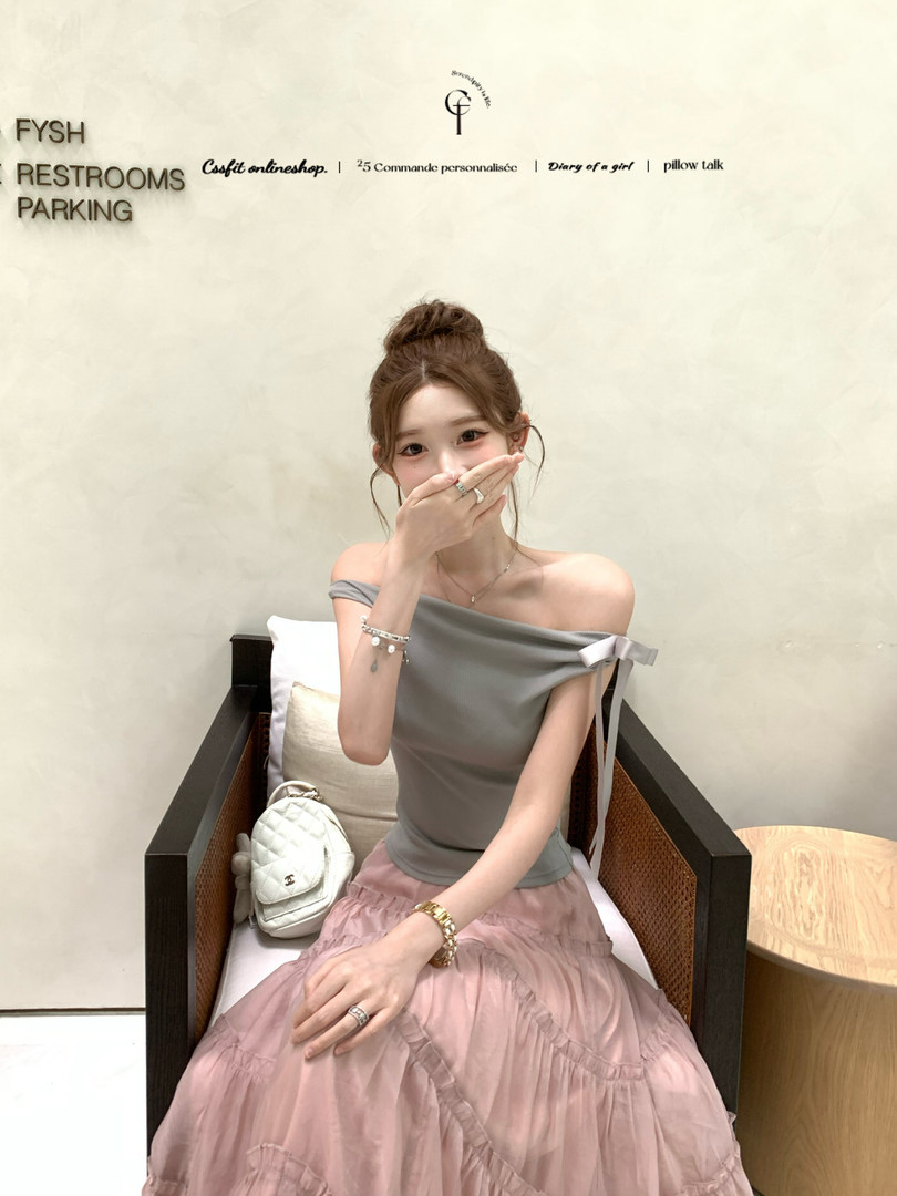 Woman wearing a gray off-shoulder top with a pink skirt, seated in an indoor setting
