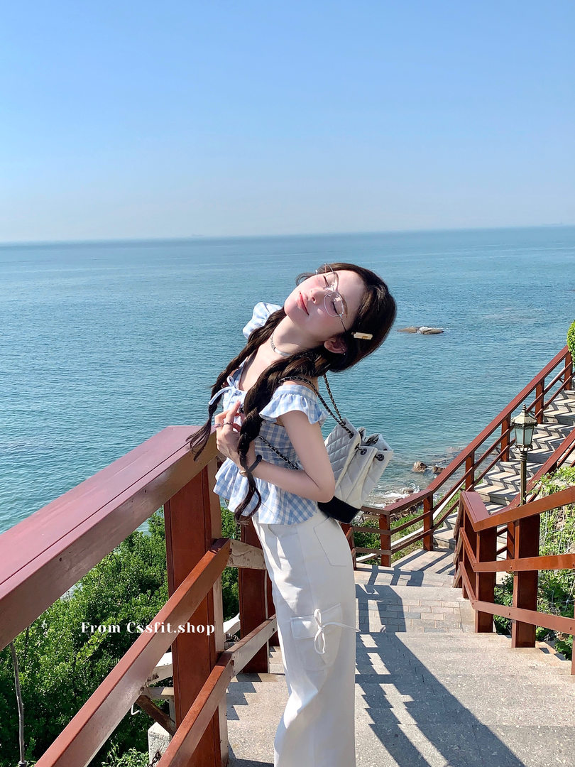 Woman wearing a blue gingham off-shoulder top with white pants by the seaside