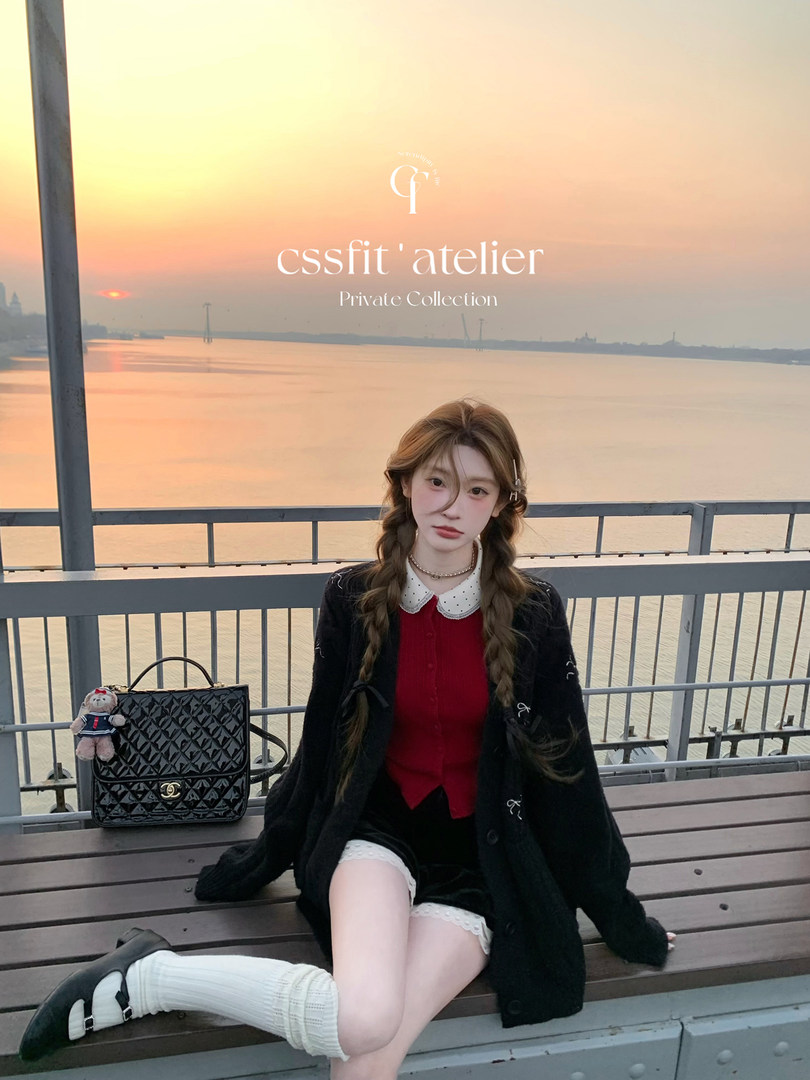 Fashion model wearing a black coat over a red top with white boots and a black handbag by the water at sunset