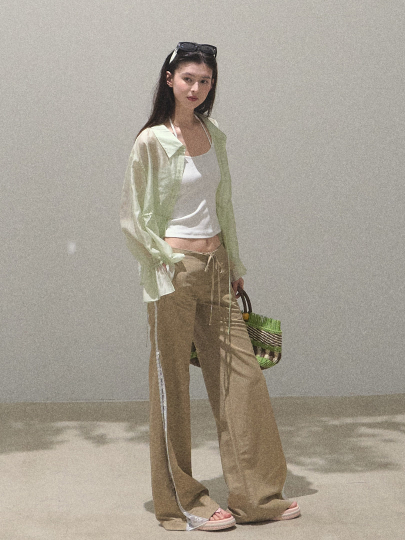Model wearing a light sheer shirt, white tank top, and wide-leg tan pants with a woven handbag