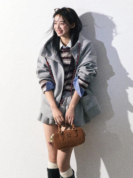 Woman wearing a gray jacket and pleated skirt while holding a brown handbag
