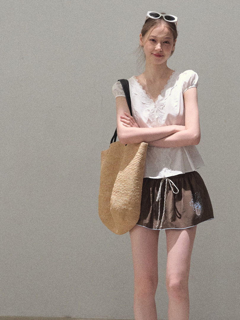 Model wearing a white lace top with a brown mini skirt and woven tote bag