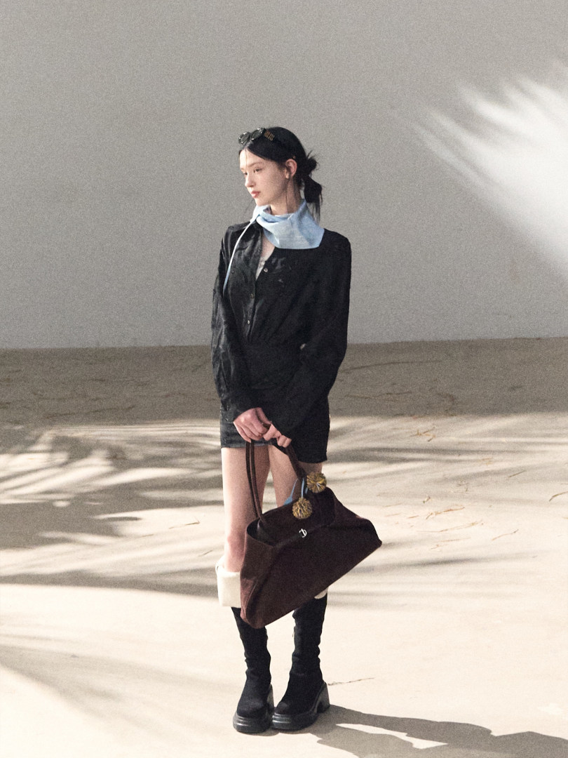 Fashion model wearing a black outfit with a light blue neck scarf and black boots, holding a dark handbag