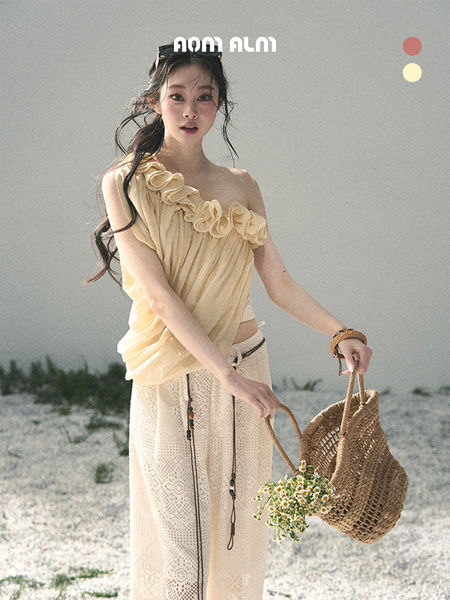 Model wearing a pale yellow one-shoulder ruffled top with a light crochet skirt and woven beach bag